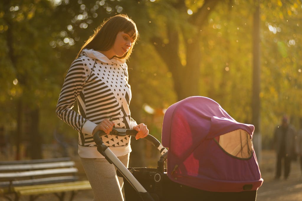 A mother walks her baby in a stroller through a sunlit park on an autumn day.