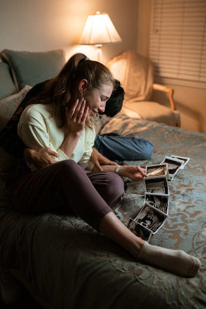 A joyful couple sits on a bed, embracing while looking at sonogram photos in a cozy bedroom.